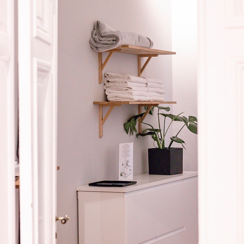 En snygg badrumshörna med rena handdukar staplade på trähyllor ovanför ett vitt skåp med en växt och bok.