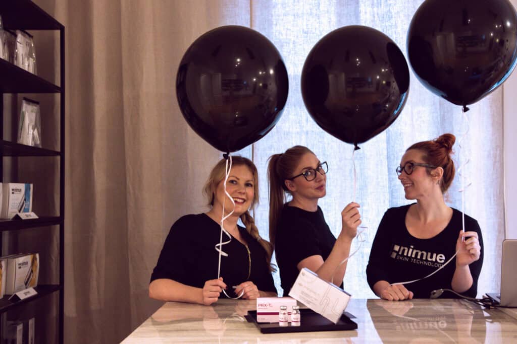 Tre leende kvinnor på ett PR-evenemang som står bakom ett skrivbord med svarta ballonger.