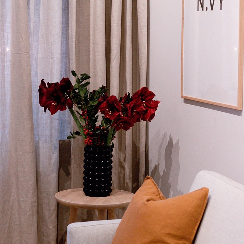 En vas med röda blommor på ett sidobord i trä i en mysig rumshörna med dekorativa kuddar och gardiner.