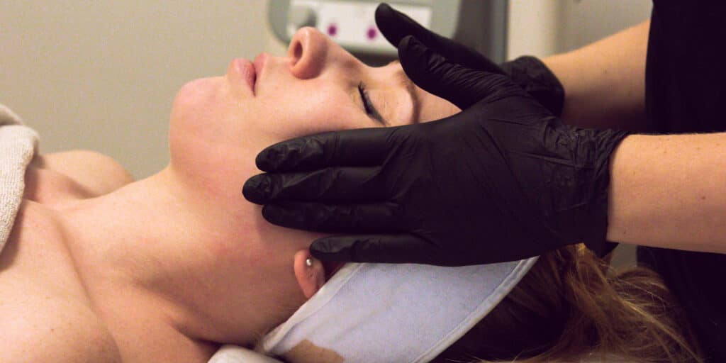 En person som får en ansiktsbehandling med en utövare som bär svarta handskar som rör vid halsen.
