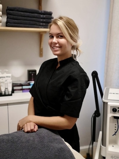 En leende kvinna i svart professionell klädsel som står bredvid ett massagebänk i ett välorganiserat behandlingsrum.