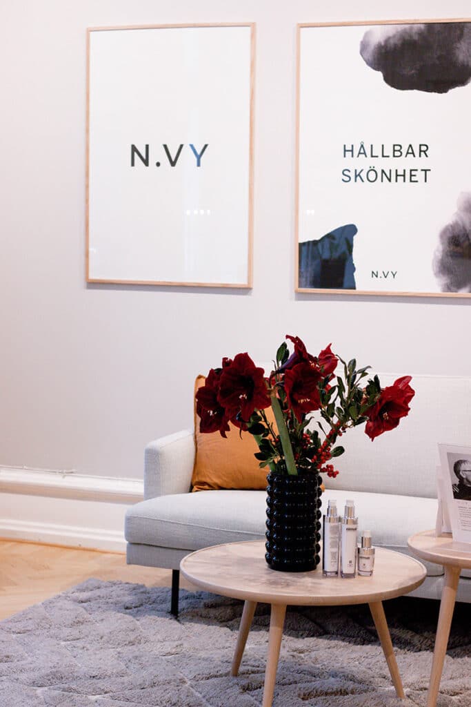 Modern vardagsrumshörna med en mysig soffa, ett runt soffbord i trä med tidningar och hudvårdsprodukter, en svart vas med röda blommor och två inramade affischer på väggen.