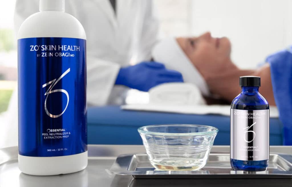 Två zo hudhälsoproduktflaskor på en bricka med en skål med vatten, i bakgrunden en vårdpersonal och en patient i en klinisk miljö.